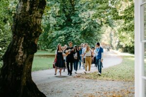A group of tour guests is led down a tree-shaded path in Hellbrunn by an entertainer with a guitar, sharing music and laughter on their way to the famous gazebo.