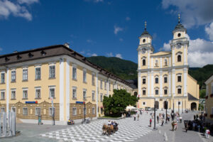 The vibrant town square of Mondsee, bustling with visitors and framed by the striking yellow façade and twin towers of the Basilica of Saint Michael, under a clear blue sky.