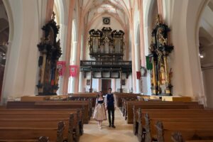 Inside a majestic church, Dana and Dmitry stand together, embodying the characters of Baron von Trapp and Maria, amidst the ornate baroque interior that fans will recognize from a famous wedding scene.