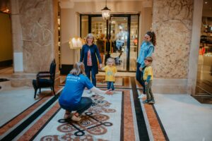 A tour guide in a blue weTours Salzburg shirt warmly reaches out to a young child in a bright yellow shirt at a luxurious hotel lobby, while another child and two adults look on, marking the beginning of a family-friendly adventure tour.