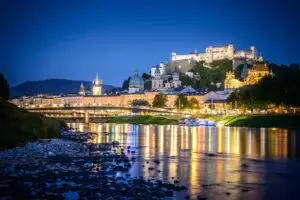 Nighttime view of Salzburg’s Old Town and fortress reflected in the Salzach River under a starry sky.